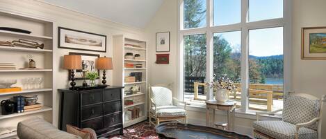 A cozy living room with a fireplace, a large window with a view of trees, and a bookshelf filled with books and decorative items.