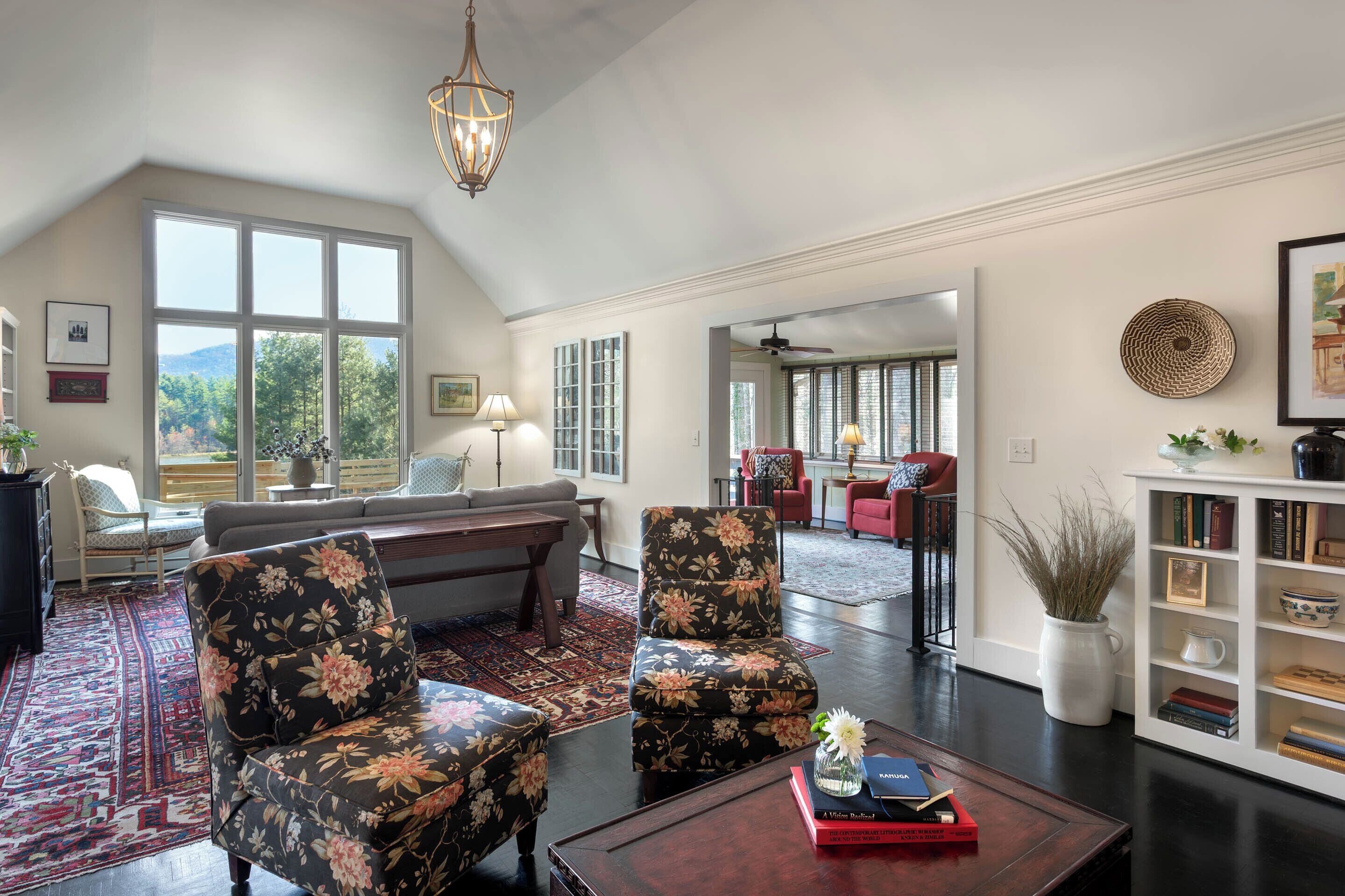 Ein geräumiges Wohnzimmer mit hohen Decken, großen Fenstern, einem dunkelholznen Couchtisch, floralen Sesseln, einer Couch und einem Bücherregal.
