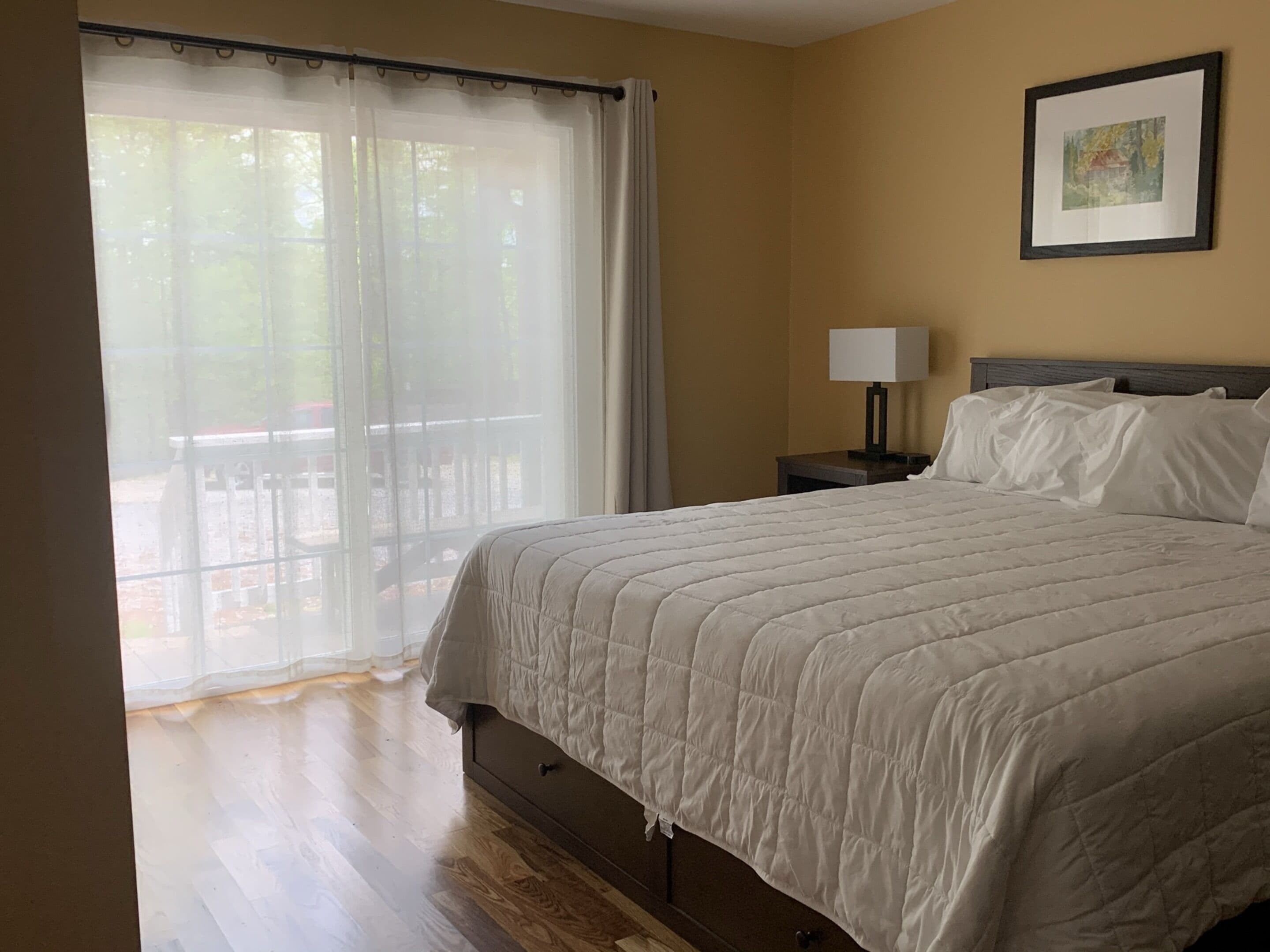 Un dormitorio con cama, una mesita de noche, una lámpara, una ventana con cortinas y un cuadro en la pared.
