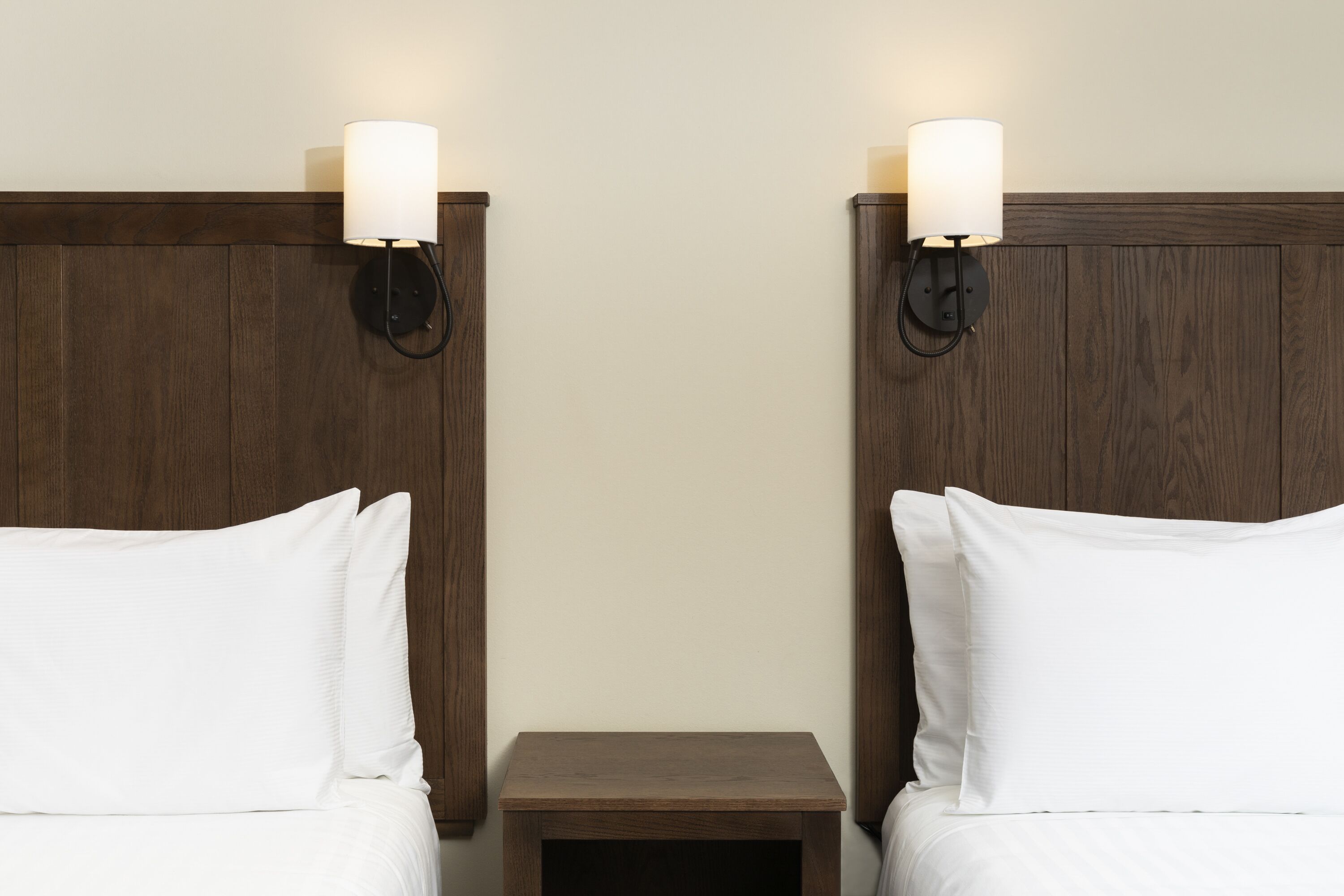 Habitación de hotel con dos camas, cada una con ropa de cama blanca, flanqueadas por cabeceros de madera y mesitas de noche fijadas a la pared.