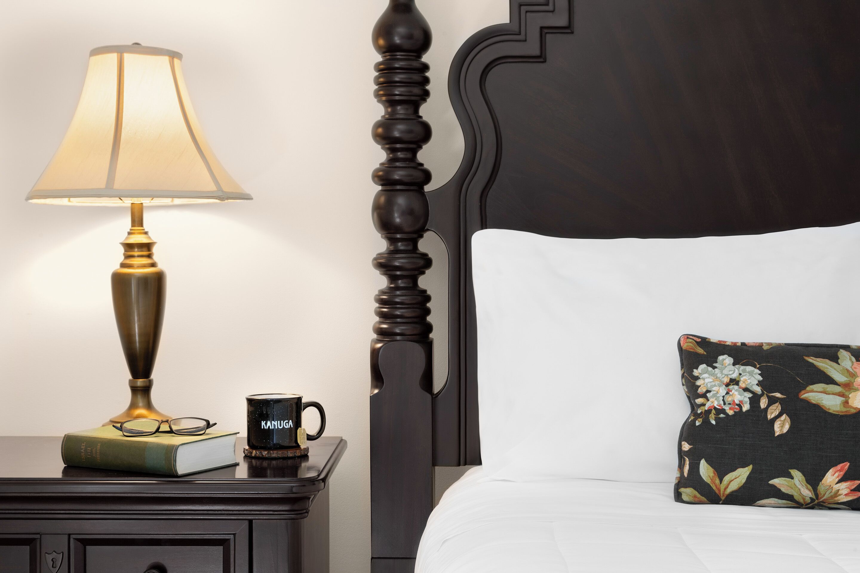A neatly made bed with a floral pillow, a bedside table with a lamp, a book, and a mug.