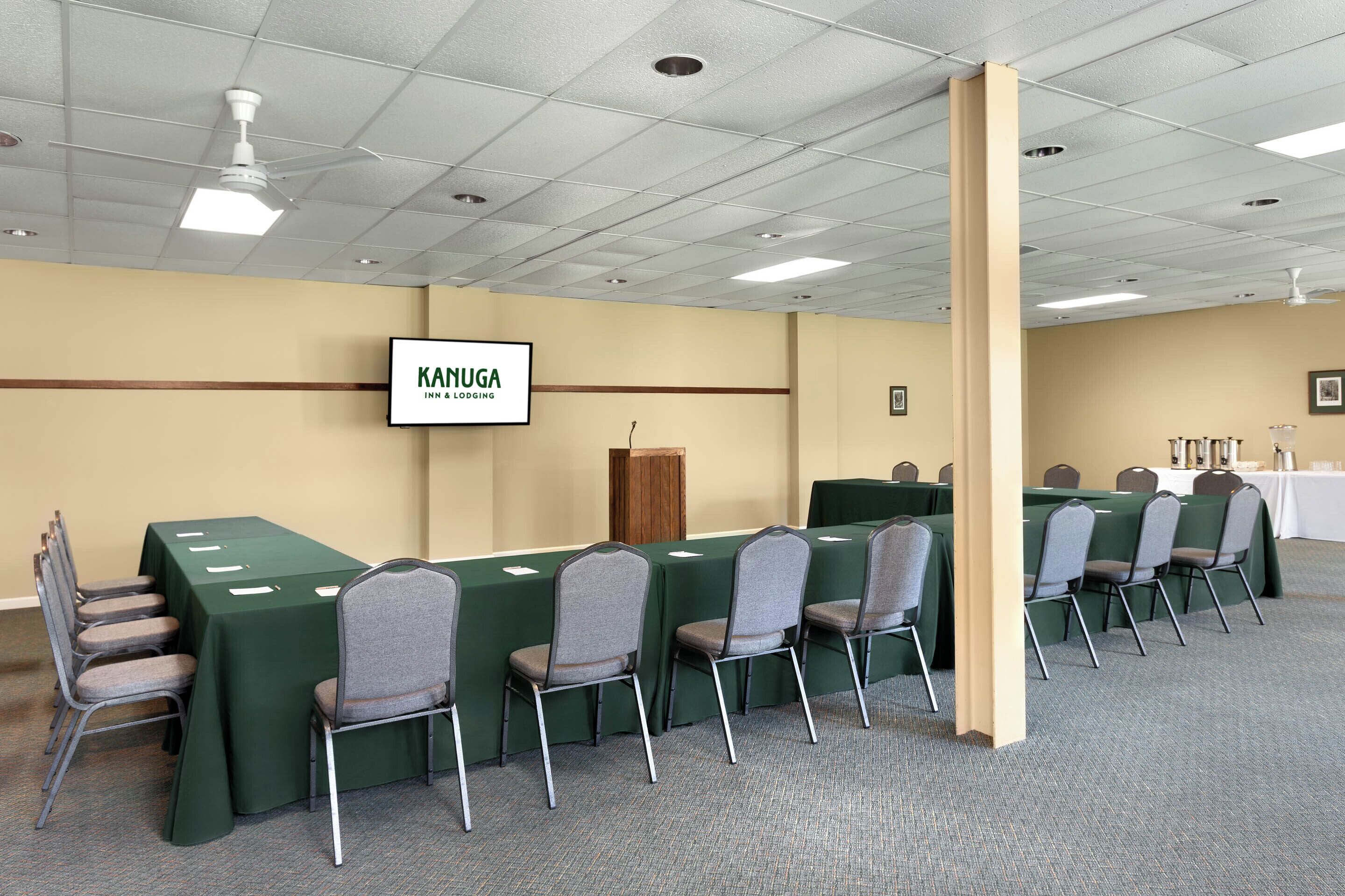 A conference room with green tables and chairs, a podium, and a sign reading "KANUGA INN & LOBOGING".