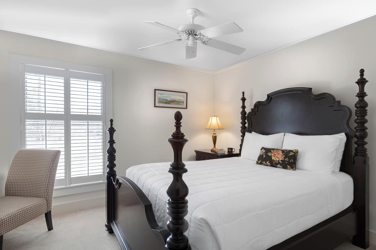 A bedroom with a four-poster bed, a chair, a nightstand with a lamp, and a ceiling fan.