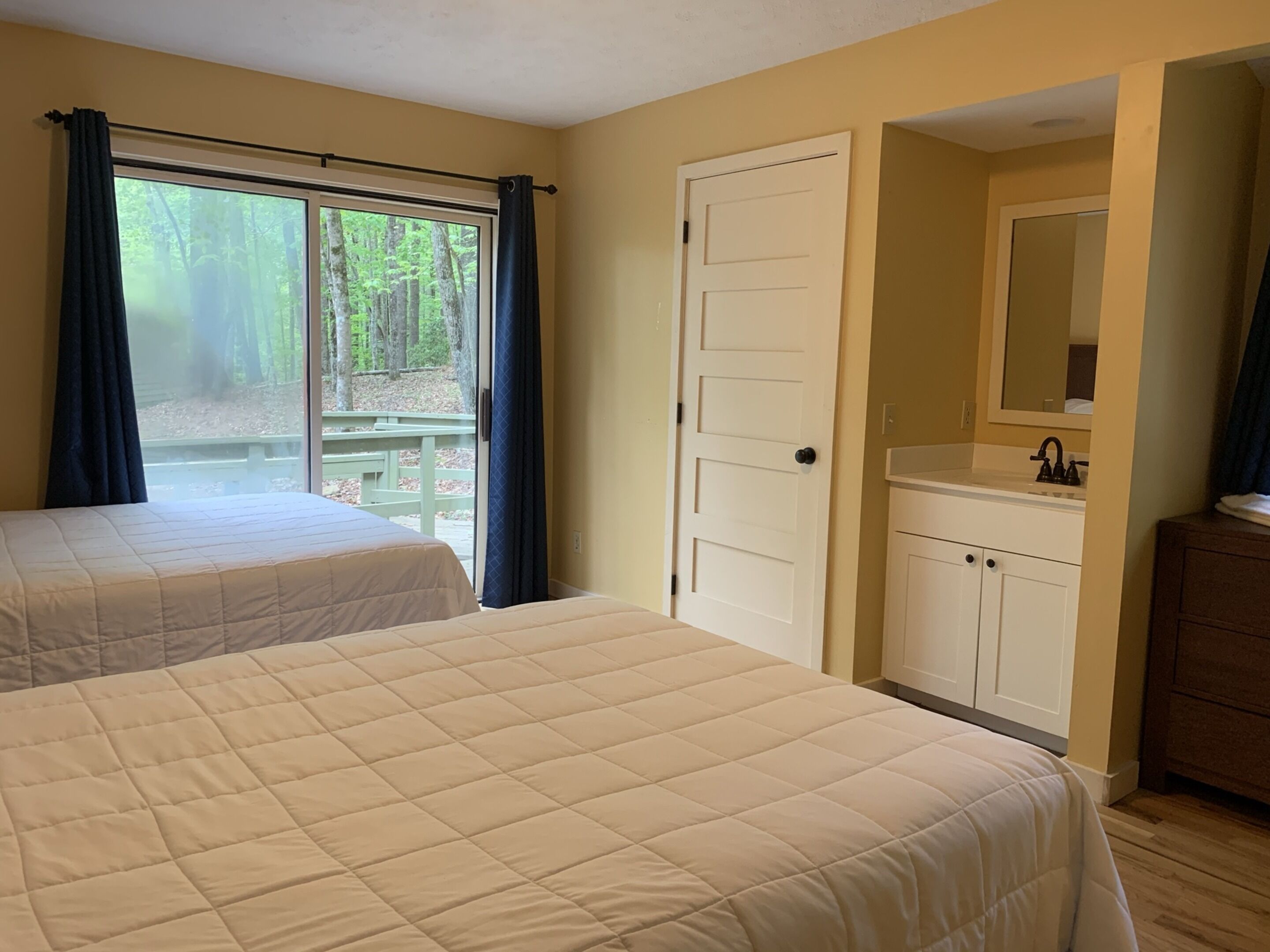 A bedroom with two beds, a window with curtains, a door, a sink, and a mirror.