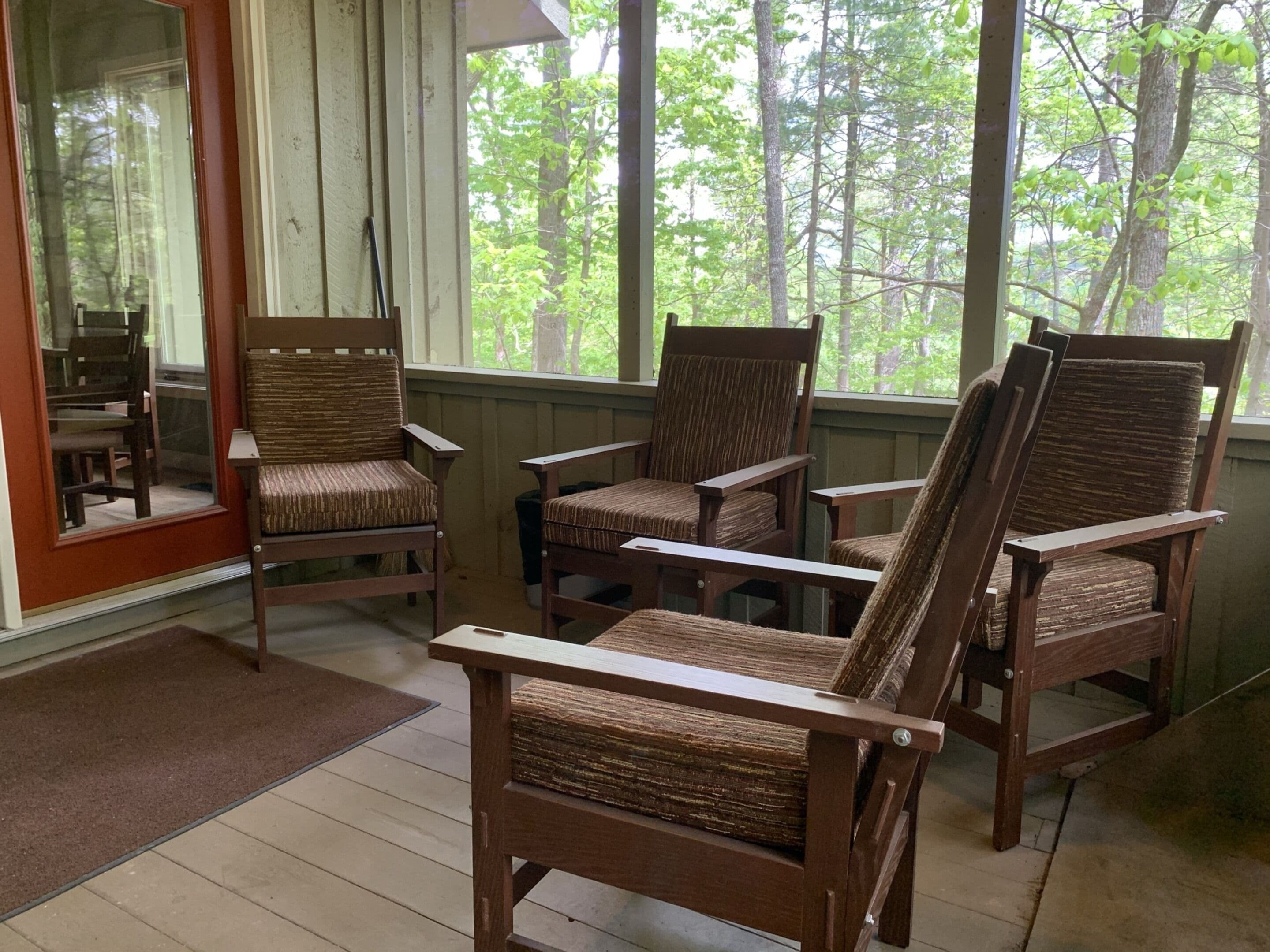 A porch with wooden chairs and a table, a glass door, and a view of trees.
