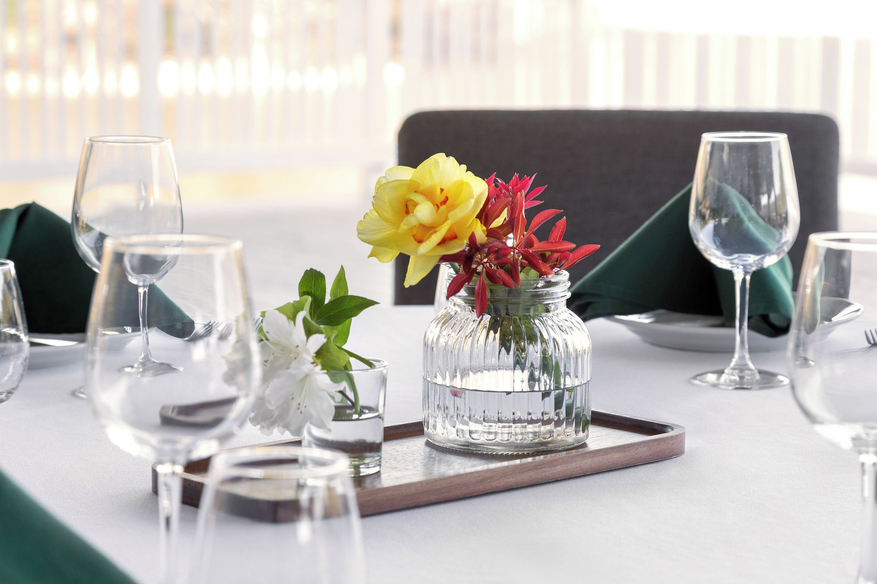 A table set for dining with a glass vase containing yellow flowers and red foliage, clear glasses, and a wooden tray.
