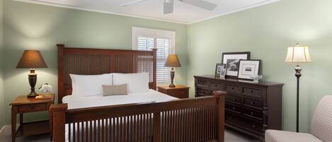 Un dormitorio con una cama de madera, dos mesitas de noche con lámparas, un aparador, una silla y un ventilador de techo.