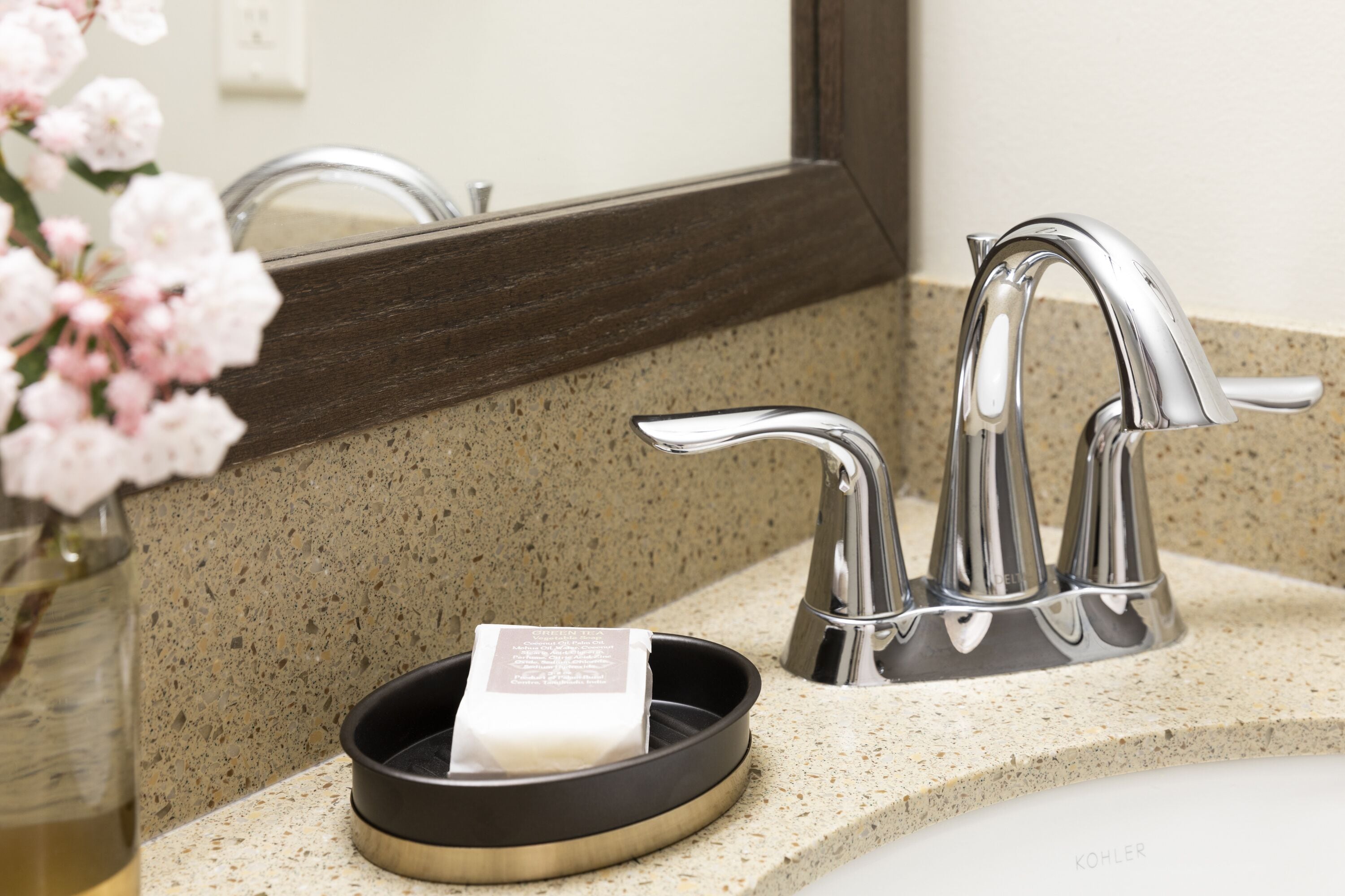 A bathroom sink with a Kohler faucet, a soap dish, and a vase of pink flowers.