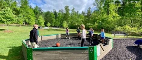 A playground with a wooden play structure surrounded by gravel and green grass, with trees in the background.