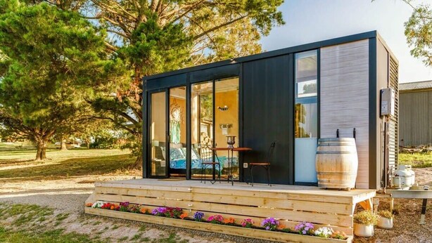 A modern, compact cabin with large glass doors, a wooden deck, and a rustic barrel.