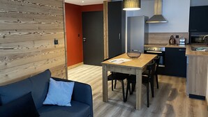 A modern kitchen with a dining area, featuring a wooden table, chairs, and a black sofa with a blue pillow. - 4-6 Pers apartment in the heart of the Chartreuse at the foot of the ski slopes (Sarcenas)