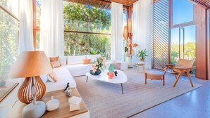 A modern living room with a sofa, coffee table, and chairs. There are large windows with curtains, a wooden ceiling, and a view of greenery outside. - PF17 Casa  05 Suítes-Praia do Forte (Mata de São João)