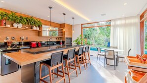 A modern kitchen with a central island, bar stools, and a dining area with a view of the outdoors. - PF17 Casa  05 Suítes-Praia do Forte (Mata de São João)