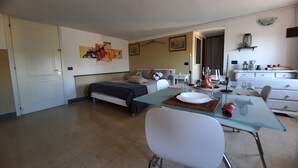 Una habitación moderna y bien iluminada con una cama, una mesa de comedor con platos y vasos, una silla blanca y una mesita de noche con una lámpara.