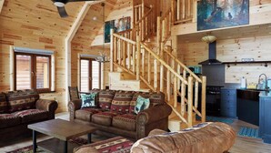 L’intérieur d’une cabane en rondins, avec un escalier en bois, un salon comprenant un ensemble de canapés, un coin repas et une cuisine.