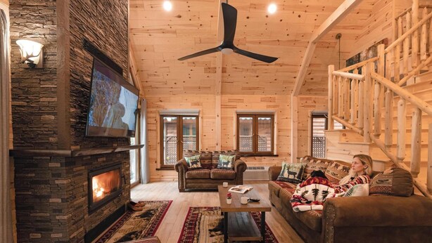 L’intérieur d’un chalet confortable, avec une cheminée en pierre, des murs en bois, un ventilateur de plafond et une grande fenêtre.