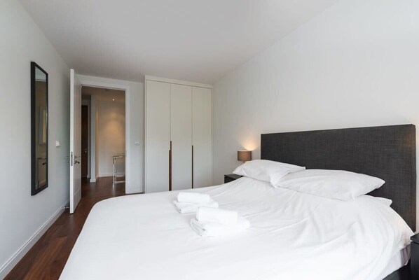 Un dormitorio moderno con una cama grande, ropa de cama blanca, un cabecero oscuro y una lámpara de noche.