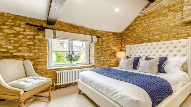 Un dormitorio con pared de piedra, una cama grande con cabecera capitonada, una silla y una ventana con vistas al verdor.