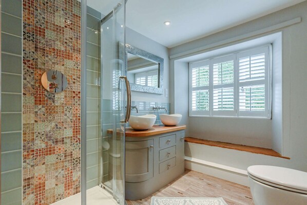 A bathroom with a glass shower enclosure, a mosaic tile wall, a wooden vanity with two sinks, a large mirror, and a window with blinds.
