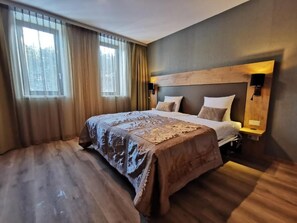 Ein modernes Hotelzimmer mit einem großen Bett, einem Holz-Kopfschild und Blick auf Bäume durch das Fenster.