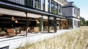 A modern building with a glass facade, outdoor seating area with chairs and tables, and a grassy landscape. - Appartementen De Egmonden (Egmond aan Zee)