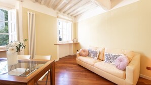 Ein Zimmer mit Holzboden, einem beigen Sofa mit floralen Kissen, einem Glastisch und einem Fenster mit Vorhängen.