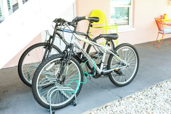 Deux vélos sont garés sur une surface en béton, à côté d’un bâtiment. On aperçoit une chaise jaune et une table à l’arrière-plan.
