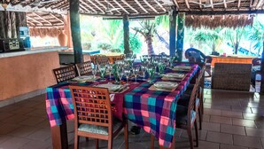 Un restaurante con una larga mesa dispuesta para comer, cubierta con un mantel colorido, sillas de madera y un techo de paja.