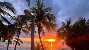 Una terraza de madera que da a una playa con palmeras y vista al atardecer.