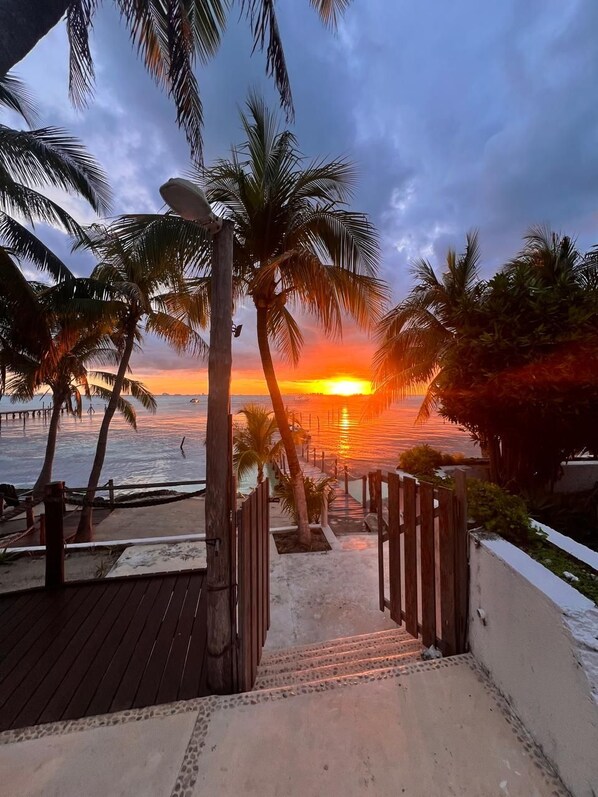 Una terraza de madera que da a una playa con palmeras y vista al atardecer.