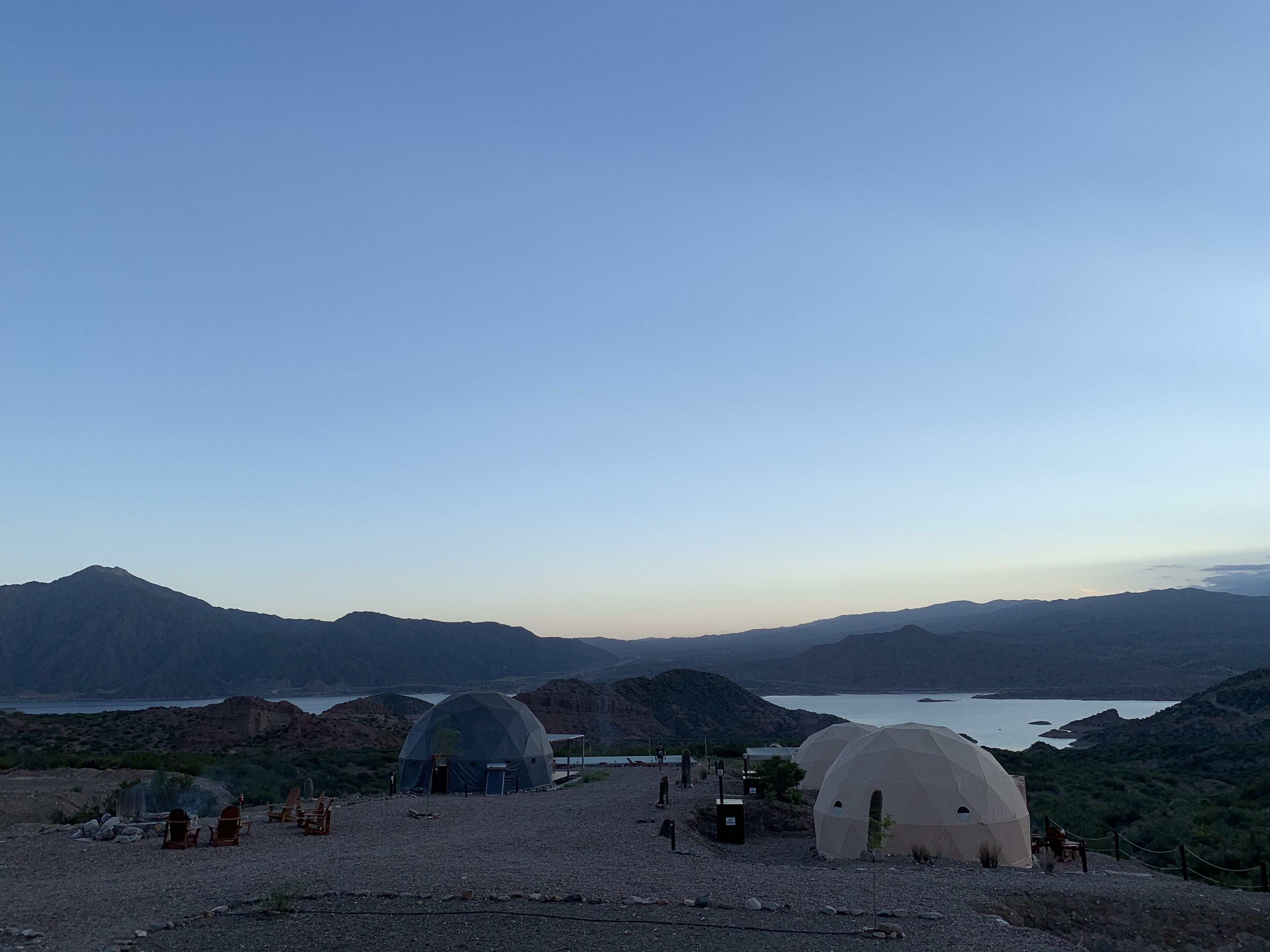 A campsite with dome-shaped tents and a few chairs, surrounded by mountains and a lake.