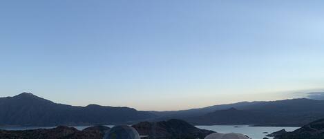 A campsite with dome-shaped tents and a few chairs, surrounded by mountains and a lake.