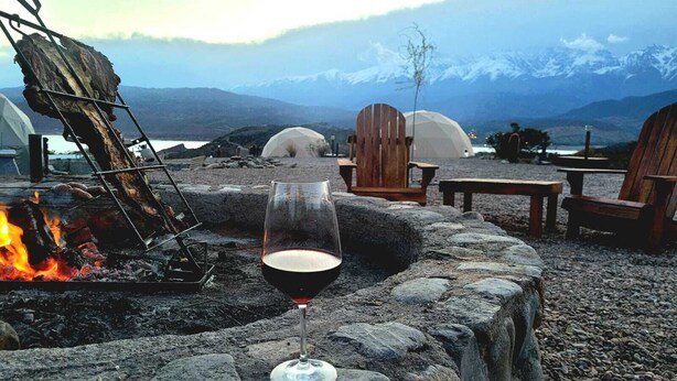 A stone fire pit with a glass of red wine, wooden chairs, and a tent in a mountainous landscape. - Denmoza Eco Lodge Glamping (Potrerillos)