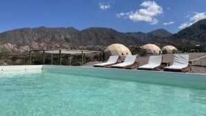 A pool with clear water, surrounded by white lounge chairs and covered umbrellas, with mountains in the background. - Denmoza Eco Lodge Glamping (Potrerillos)