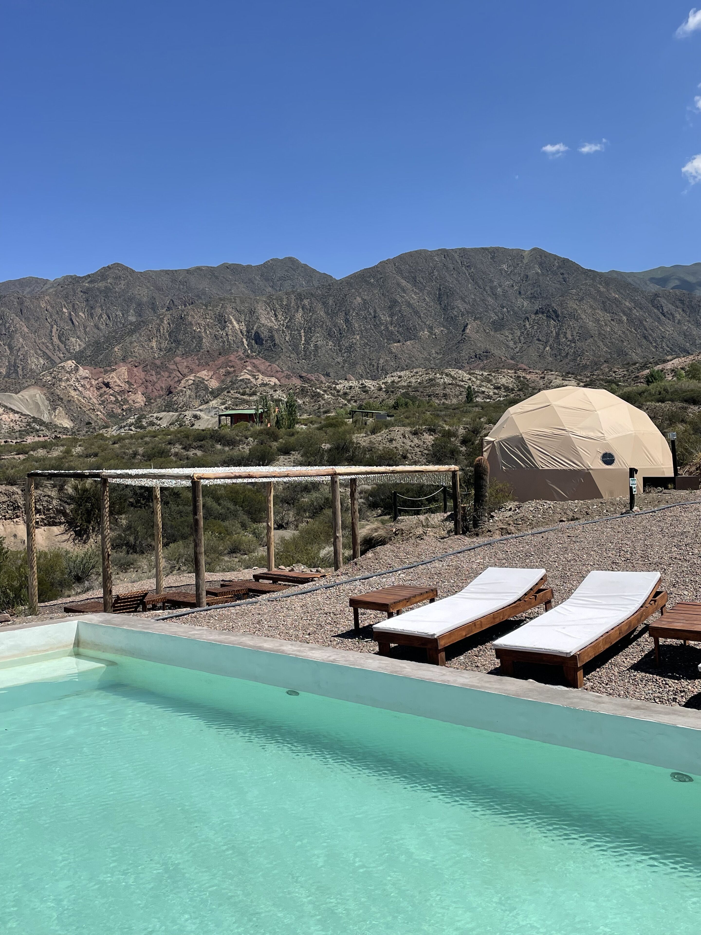 A pool area with lounge chairs and a pergola, a dome structure, and a mountainous backdrop.