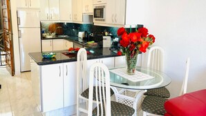 Cozinha moderna com armários brancos, bancada preta e mesa de jantar de vidro com cadeiras brancas.