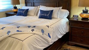 A neatly made bed with white bedding and blue pillows, a wooden headboard, a bedside table with a lamp, and a digital clock. - Ellie’s Loft in the heart of downtown Nashville, IN. (Nashville)