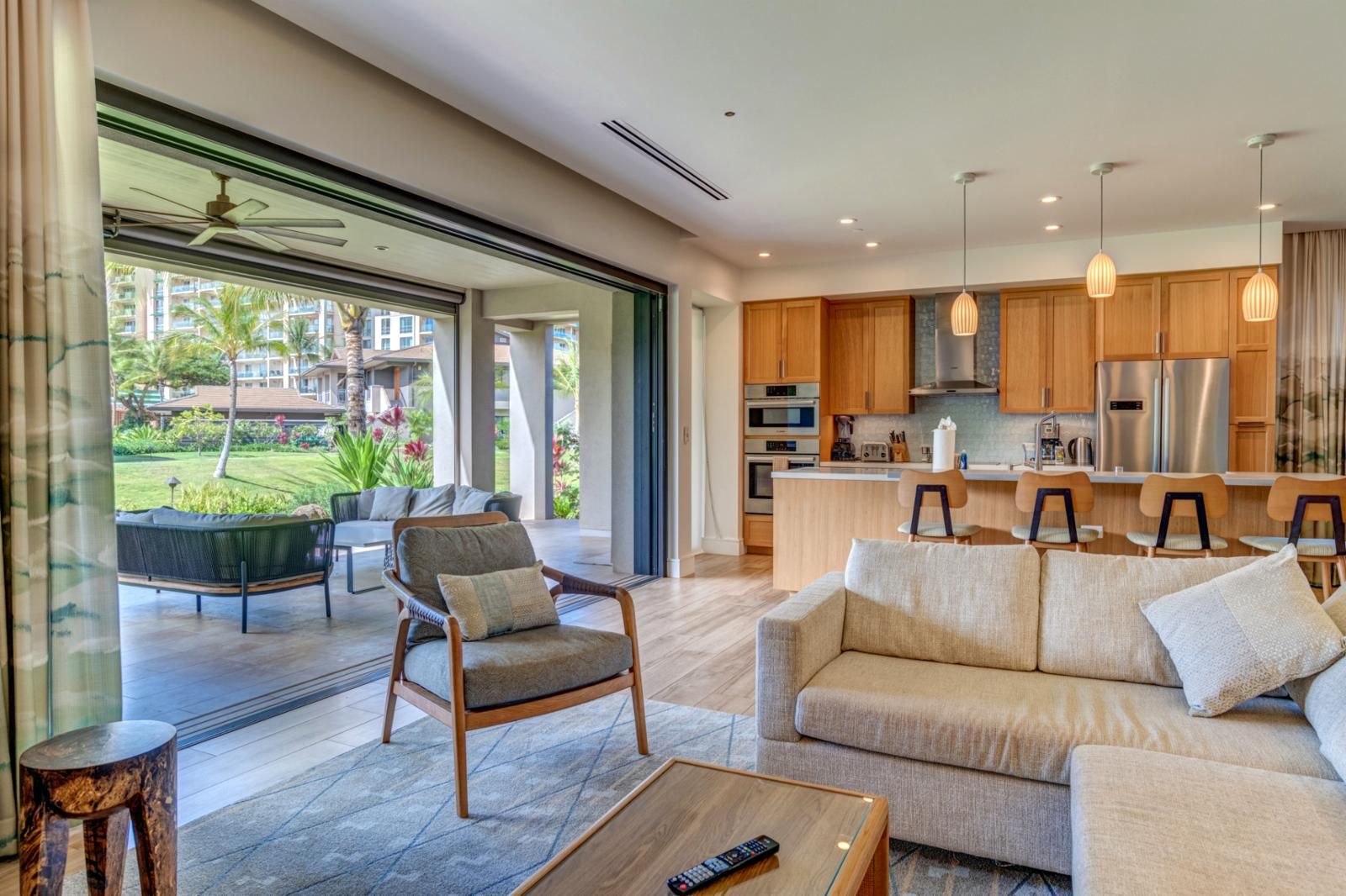 A modern living room with a sofa, armchair, and coffee table. A kitchen with wooden cabinets and stainless steel appliances is visible in the background.