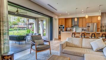A modern living room with a sofa, armchair, and coffee table. A kitchen with wooden cabinets and stainless steel appliances is visible in the background.