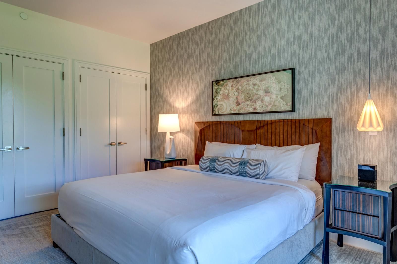 A hotel room with a large bed, a wooden headboard, a nightstand with a lamp, and a framed picture on the wall.