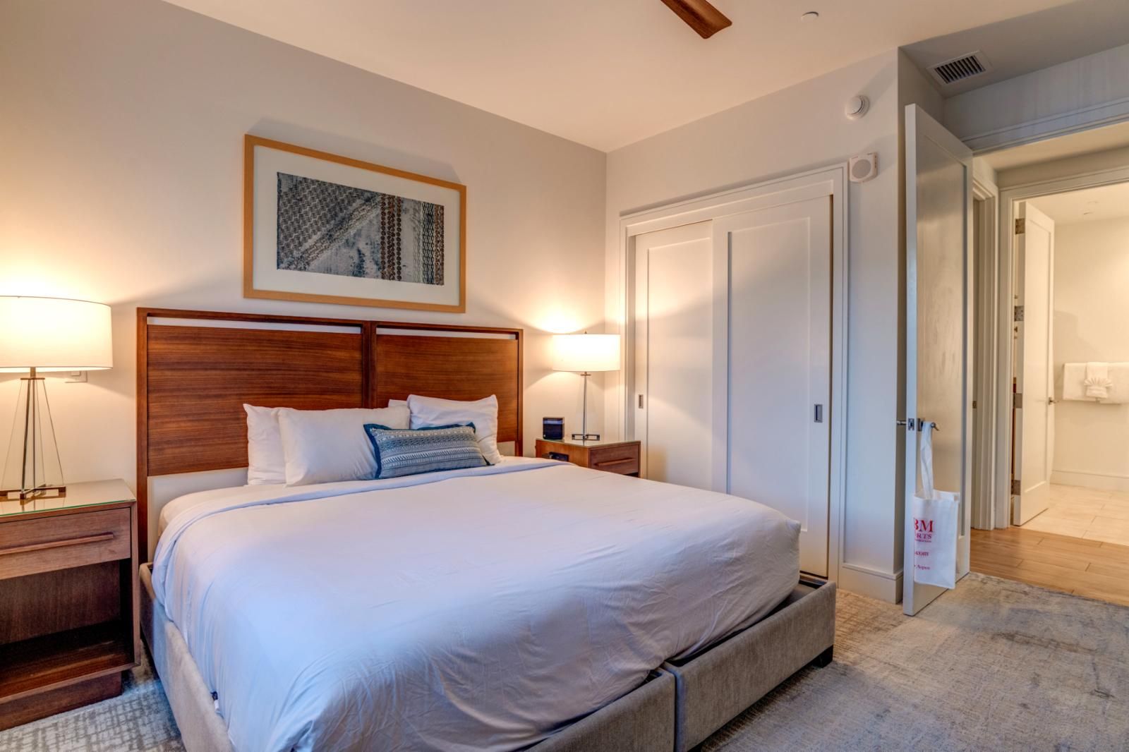 A neatly made bed with a wooden headboard, two bedside tables with lamps, a framed artwork above the bed, and a door leading to another room.