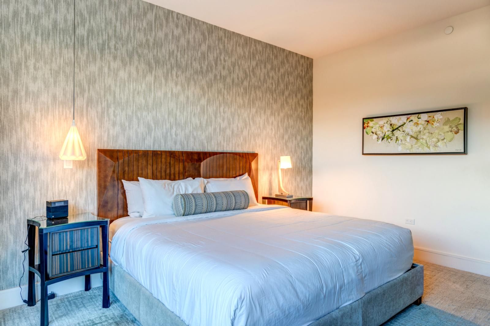 A modern hotel room with a large bed, wooden headboard, two bedside tables with lamps, a small side table, and a framed floral artwork on the wall.