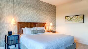 A modern hotel room with a large bed, wooden headboard, two bedside tables with lamps, a small side table, and a framed floral artwork on the wall.