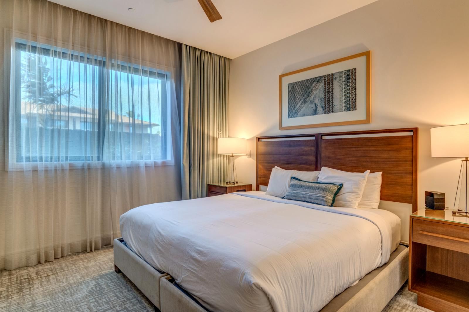 Une chambre d’hôtel avec un grand lit, une tête de lit, une table de chevet, une lampe et une fenêtre avec des rideaux.