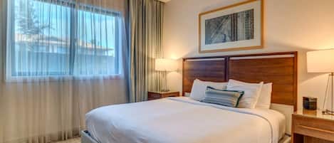 Une chambre d’hôtel avec un grand lit, une tête de lit, une table de chevet, une lampe et une fenêtre avec des rideaux.