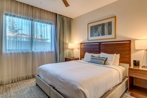 Quarto de hotel com uma cama grande, cabeceira, mesa de cabeceira, luminária e janela com cortinas.