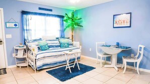 Un dormitorio con cama, escritorio, sillas, una mesita, una planta y una ventana con cortinas.