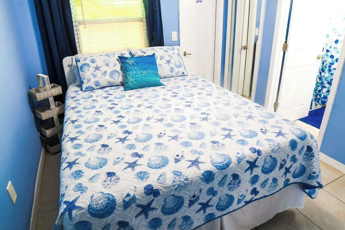 A bedroom with a bed covered in a blue and white patterned bedspread, a blue pillow, and a small cross-shaped decoration on the nightstand.