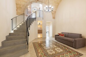 Una escalera con barandal negro que conduce a una plataforma, un sofá de cuero marrón con cojines rojos, y una alfombra estampada en el suelo.