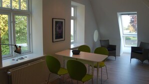 Een eethoek met een witte tafel en groene stoelen, grote ramen en een lijst met een foto aan de muur.
